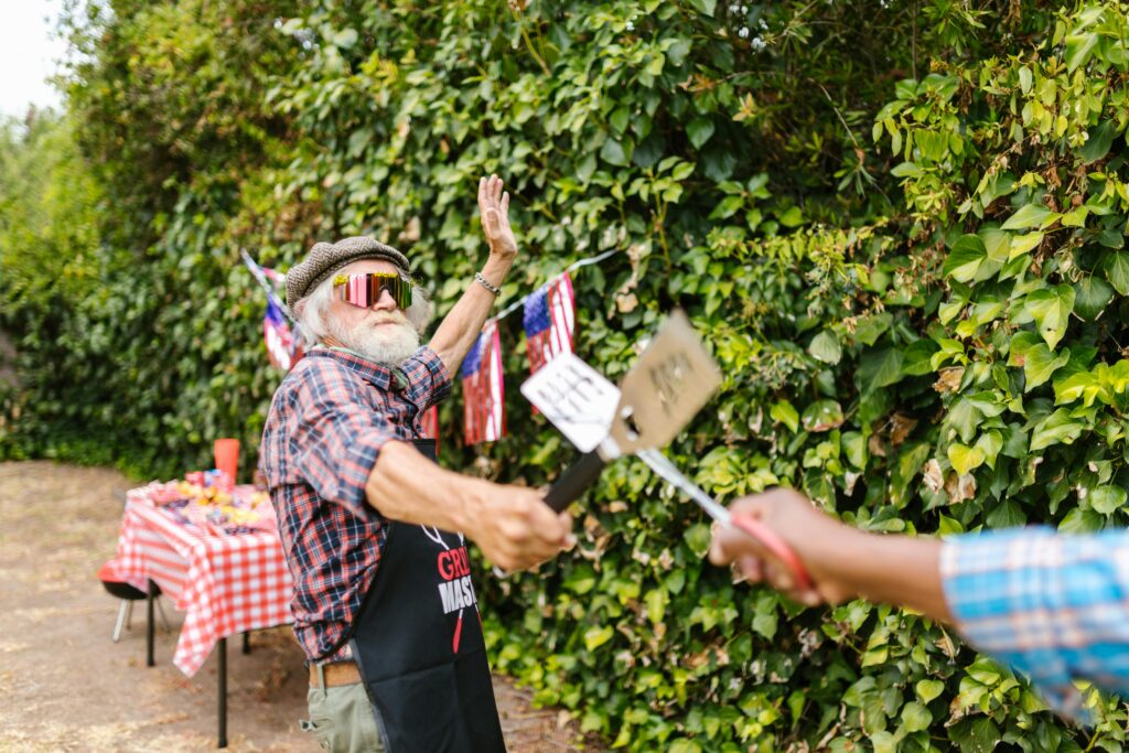 senior man plays sword fighting with spatulas at a bbq with man off camera